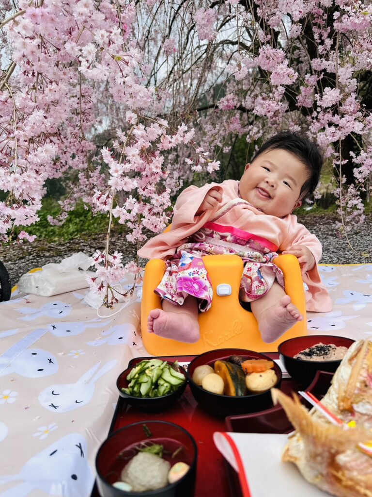お食い始めを桜の下で。花も笑顔も春らんまん。満開の花咲く素敵な人生を送ってね